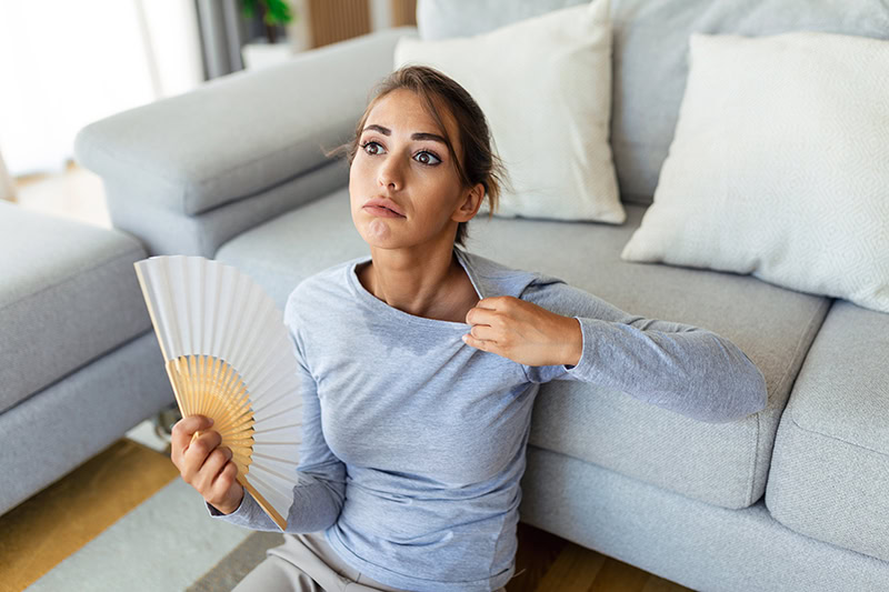 A young woman sits on the floor, leaning against a gray couch, looking visibly exhausted and overheated.