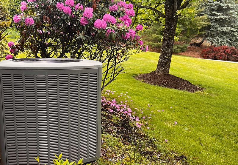 A large gray outdoor air conditioning condenser unit stands on the edge of a meticulously landscaped yard. Positioned directly behind the unit is a vibrant green bush covered in blooming pink rhododendron flowers.