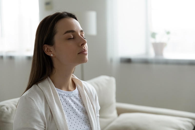 Why Do I Need a Humidifier? Woman comfortably sitting on a grey couch with her arms behind her head in a resting position.