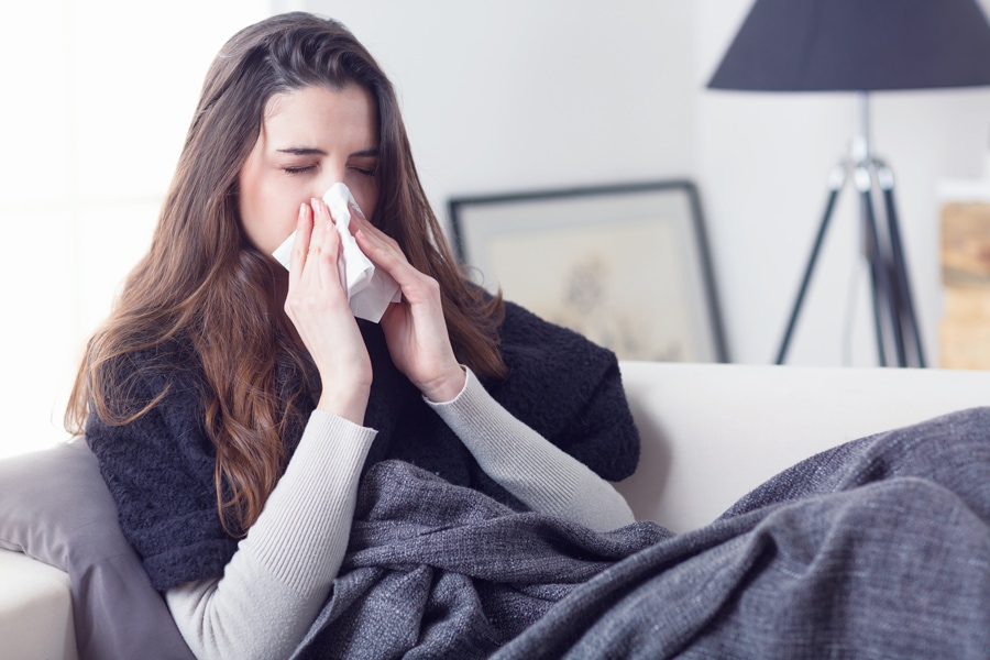 Young attractive woman lying sick at home couch in cold and flu sneezing nose with tissue covering with blanket, What Does Indoor Air Quality Have to Do With Allergies?