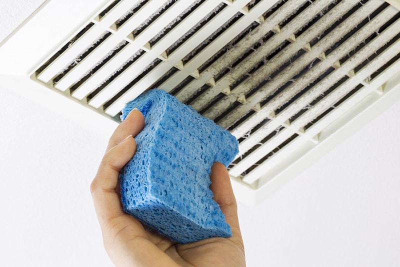 Why Air Duct Cleaning Is Important. Close up horizontal photo of female hand cleaning dirty bathroom fan vent cover with blue sponge.